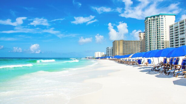 Ein Strand mit weißem Sand, klarem türkisblauem Wasser und blauen Sonnenschirmen.