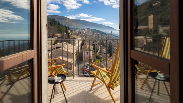 A balcony with a view of a historic town, wooden chairs, and a small table with a bowl of food.