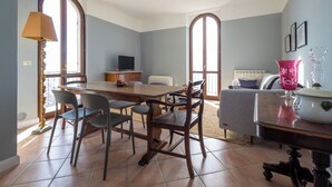 A dining area with a wooden table and chairs, a television, and a sofa in a room with large windows and a radiator.