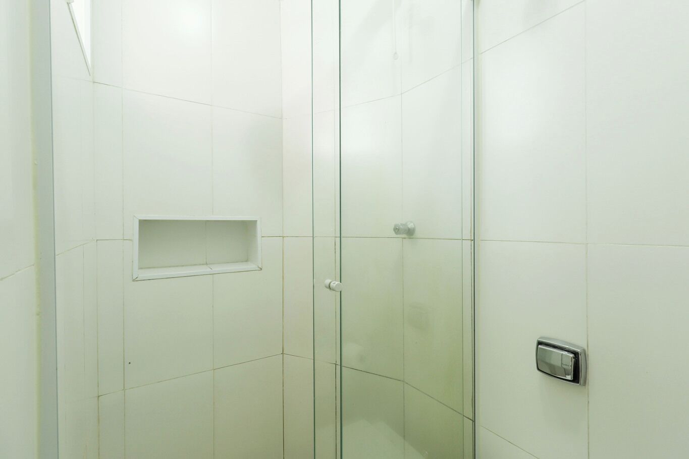 A bathroom with white tiled walls, a glass shower enclosure, a small shelf, and a thermostat on the wall.