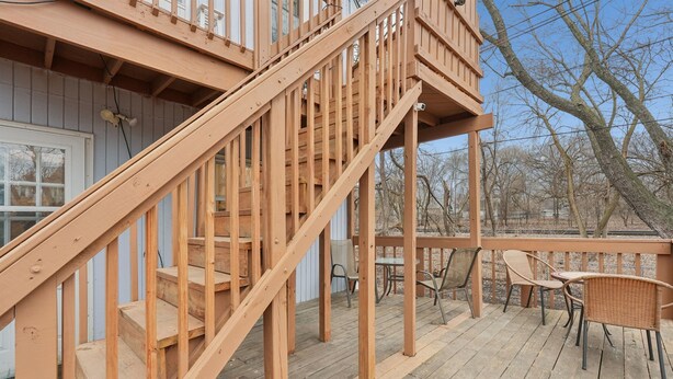Um deck de madeira com escadas que levam a um segundo andar, móveis de exterior e vista para árvores.