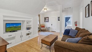 Uma sala de estar com televisão de tela plana, armários brancos, um sofá marrom e uma mesa de centro de madeira.