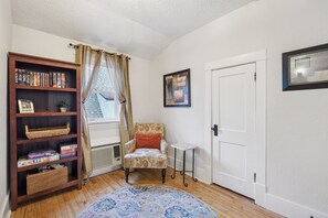 Ein Zimmer mit einer Holzbibliothek, einem gemusterten Sessel, einem kleinen Tisch, einer Tür und einem gerahmten Bild an der Wand.