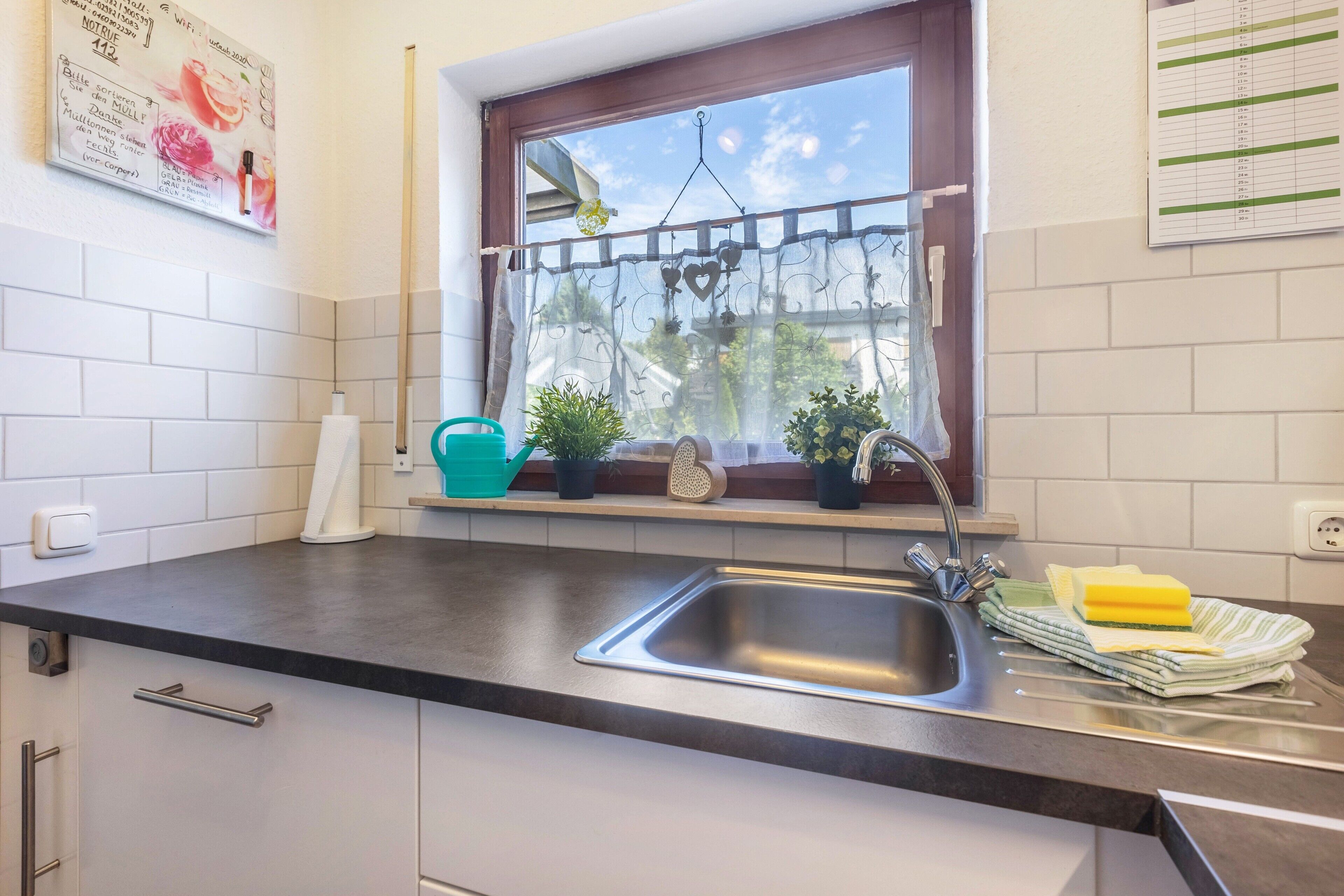 A kitchen with a sink, a window with a curtain, a calendar, and a potted plant.