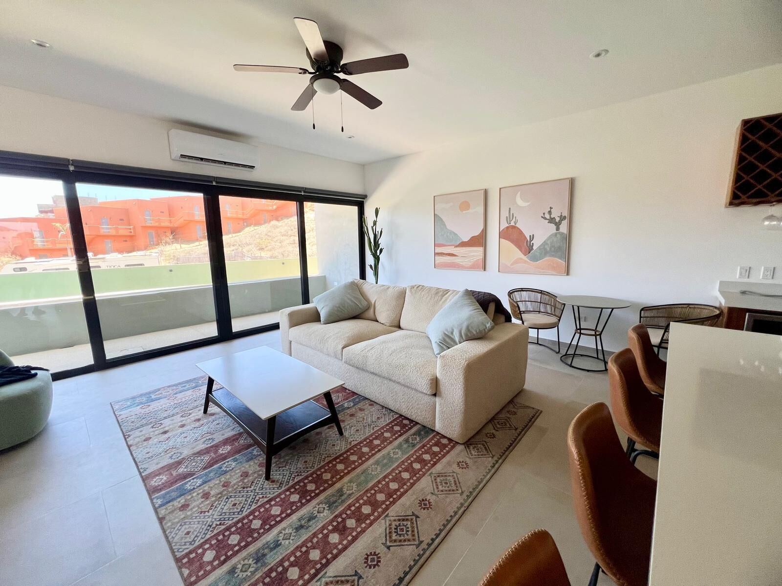 A spacious living room with a sofa, coffee table, and chairs. There is a ceiling fan, large windows, and artwork on the walls.