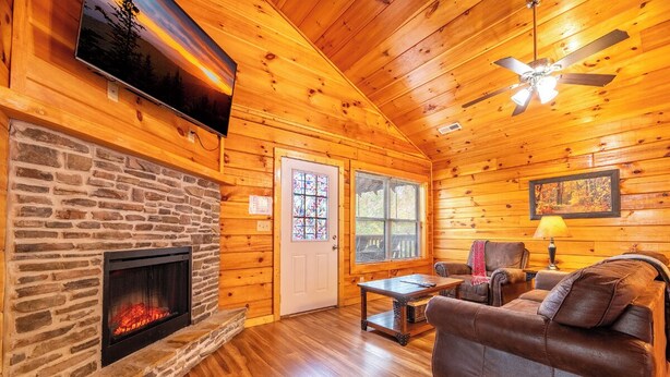 A cozy cabin interior with a stone fireplace, wooden walls, a ceiling fan, and a comfortable seating area. - *2BD w/ POOL Access Near TOWN! Theater + Hot Tub* (Pigeon Forge)