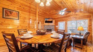 A wooden cabin interior with a round dining table, chairs, a fireplace, a television, and a ceiling fan. - *2BD w/ POOL Access Near TOWN! Theater + Hot Tub* (Pigeon Forge)