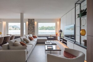 Ein modernes Wohnzimmer mit einer Couch, einem Couchtisch und einem Fernseher. Große Fenster bieten Blick auf das Meer und die Berge.