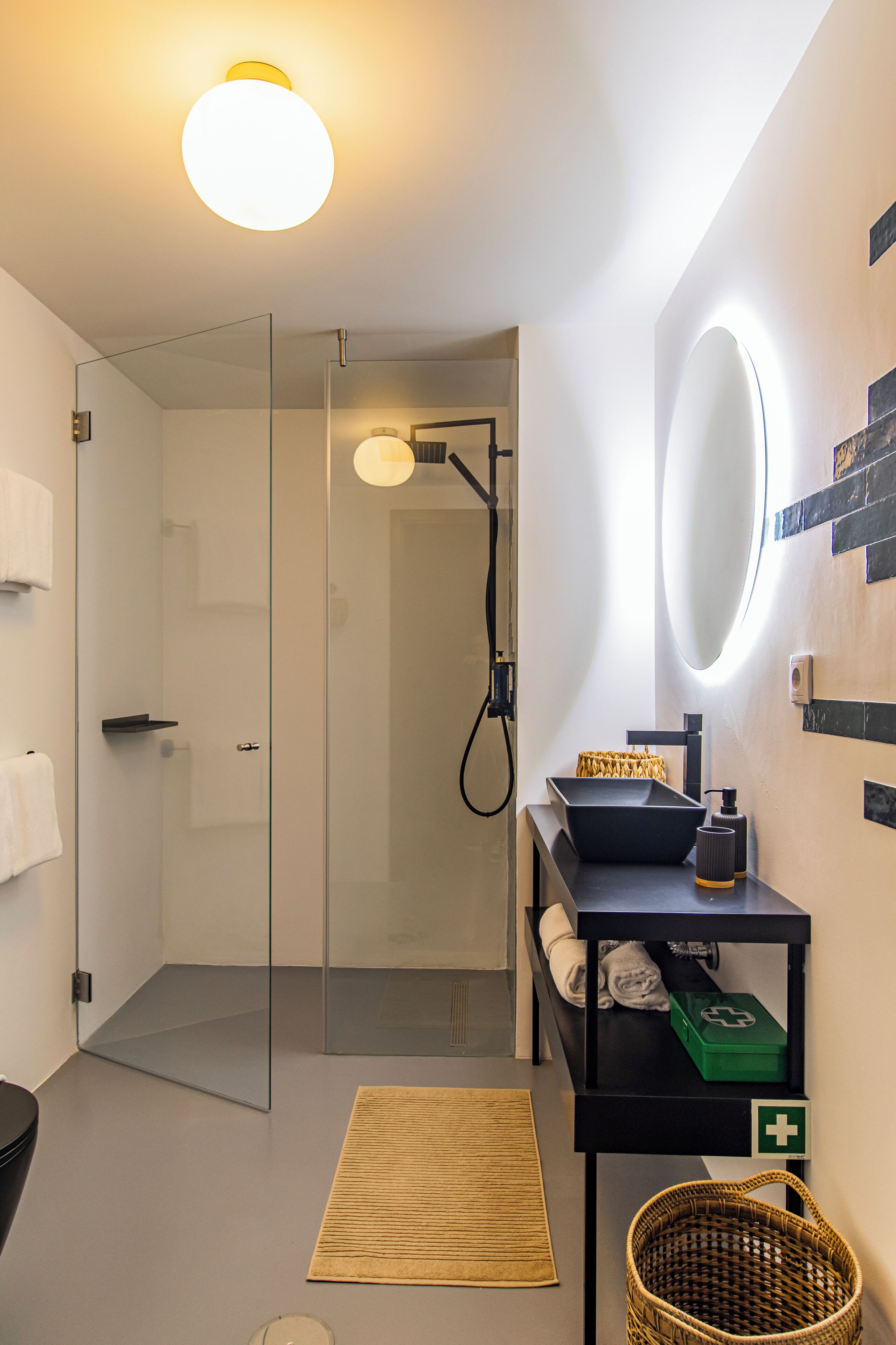 Een moderne badkamer met een glazen douchecabine, een zwart wastafelmeubel met een komvormige wasbak, een ronde spiegel en een kleine, groene verbanddoos op een zwart plankje.