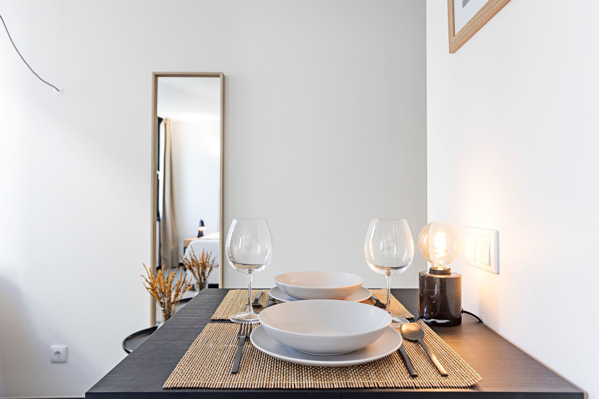 Una mesa de comedor dispuesta con platos, cubiertos y copas de vino, con una lámpara y un espejo al fondo.