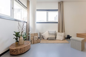 A modern living room with a large window, a wooden coffee table, a sofa with patterned cushions, and a small ottoman.