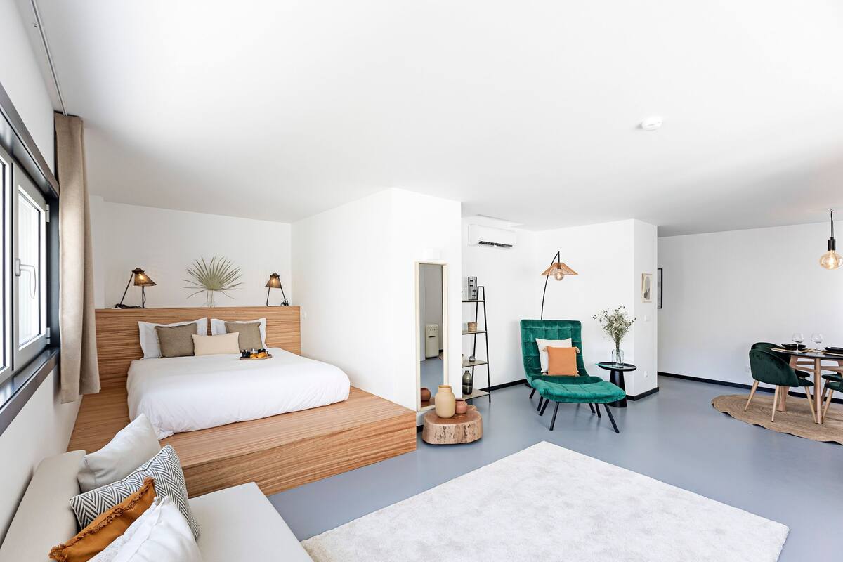 Una habitación de hotel moderna con una cama de madera, un sillón verde, un rincón de comedor y un ventanal grande.