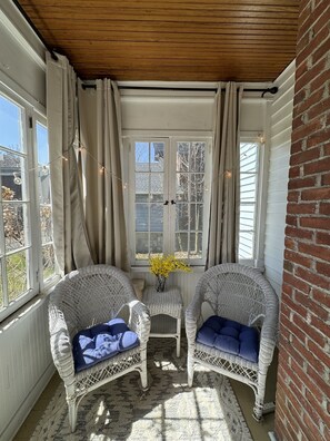 A cozy room with two wicker chairs, a small table with a vase of flowers, and a view through a window with curtains.