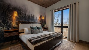 Ein Schlafzimmer mit einem großen Bett, einer Waldlandschaftsmalerei, zwei Nachttischlampen und Blick durch ein Fenster.