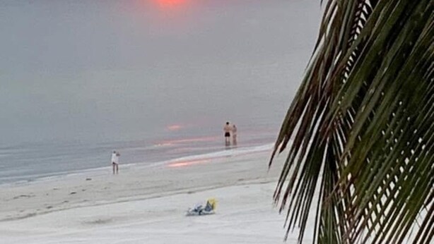Strandszene bei Sonnenuntergang, mit Palmen und ein paar Personen.