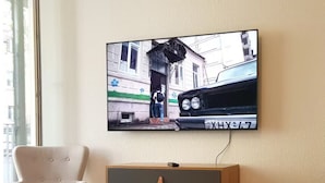 A room with a television mounted on the wall displaying a street scene, a wooden dresser with drawers, a light-colored armchair, and a bed with a white duvet.
