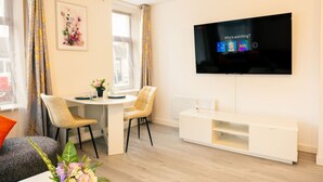 A modern living room with a flat-screen TV mounted on the wall, a round dining table with chairs, a white entertainment unit, and a window with curtains. - Stylish 3-Bed Flat Near Cardiff City Centre & Uni (Cardiff)