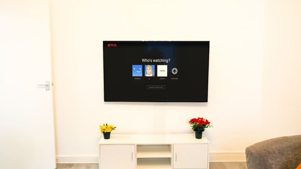 A modern living room with a flat-screen TV mounted on the wall, a white entertainment unit below, and a door to the left. - Cozy 2BR Flat Near Cardiff City Centre | Stadium| Shops | Uni (Cardiff)