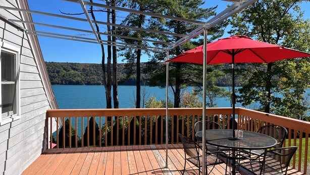 Una terraza de madera con una mesa, sillas, un paraguas rojo y vistas a un lago y árboles.