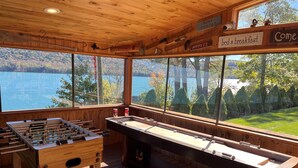 Une pièce au plancher de bois, équipée d’un baby-foot et d’une table de billard, avec de grandes fenêtres donnant sur un lac et des arbres.