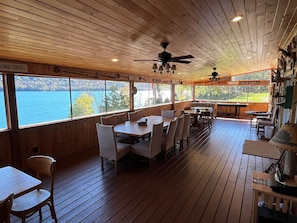 A wooden deck with tables and chairs, large windows overlooking a lake, and a ceiling fan.