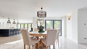 A modern dining area with a round table, four chairs, and a chandelier. - Mayrose House: 6BR 4BATH Luxe Spacious Retreat (Takeley)