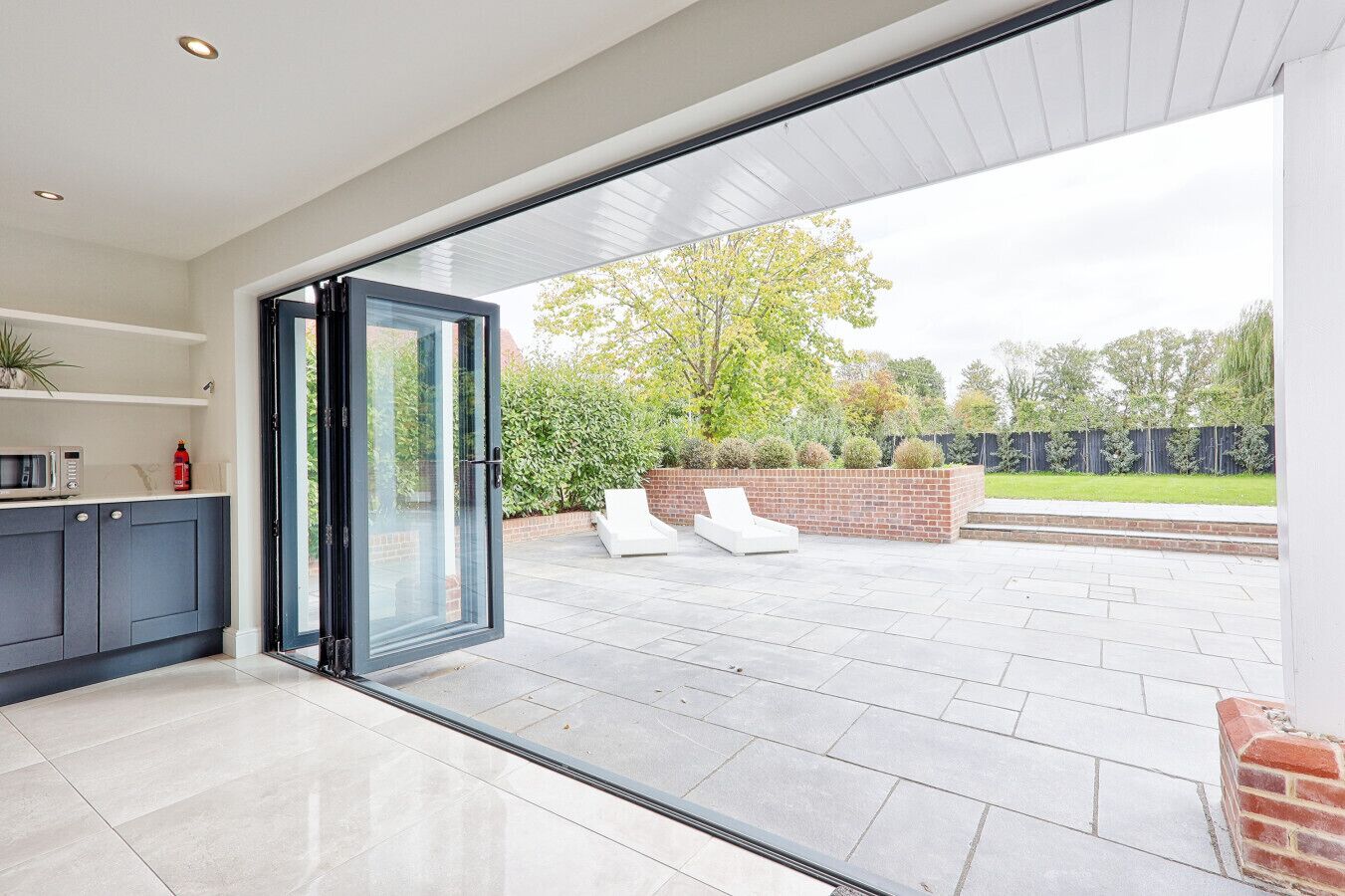 Une cuisine moderne avec des meubles sombres et une porte vitrée donnant sur un patio agrémenté d’un mur en briques, de chaises longues blanches et offrant une vue sur un jardin.