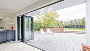 A modern kitchen with dark cabinets and a glass door leading to a patio with a brick wall, white lounge chairs, and a view of a garden. - Mayrose House: 6BR 4BATH Luxe Spacious Retreat (Takeley)