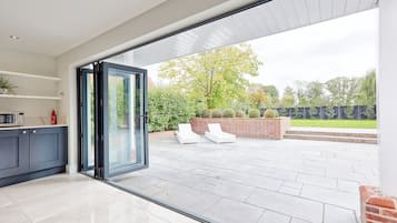 Une cuisine moderne avec des meubles sombres et une porte vitrée donnant sur un patio agrémenté d’un mur en briques, de chaises longues blanches et offrant une vue sur un jardin.
