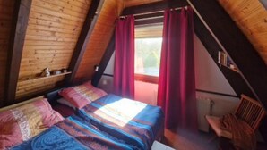 Una camera da letto con soffitto spiovente, travi in legno, un letto con lenzuola decorate, una sedia e una finestra con tende rosse.