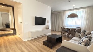 Een moderne woonkamer met een flatscreen televisie aan de muur, een bank, een salontafel en een eethoek met een tafel en stoelen.