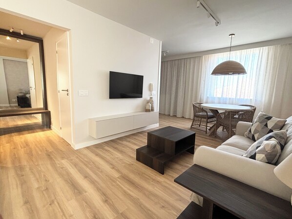 A modern living room with a flat-screen TV mounted on the wall, a sofa, a coffee table, and a dining area with a table and chairs. - Studio with a Pool in Downtown Santa Cruz (SANTA CRUZ DE TENERIFE)