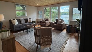 Amplia sala de estar con techo de madera, ventanas amplias con vistas a un lago y un rincón acogedor con sofá, sillones y alfombra estampada.