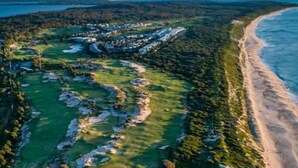 Vue aérienne d’un parcours de golf situé sur la côte, avec une plage et une forêt dense.