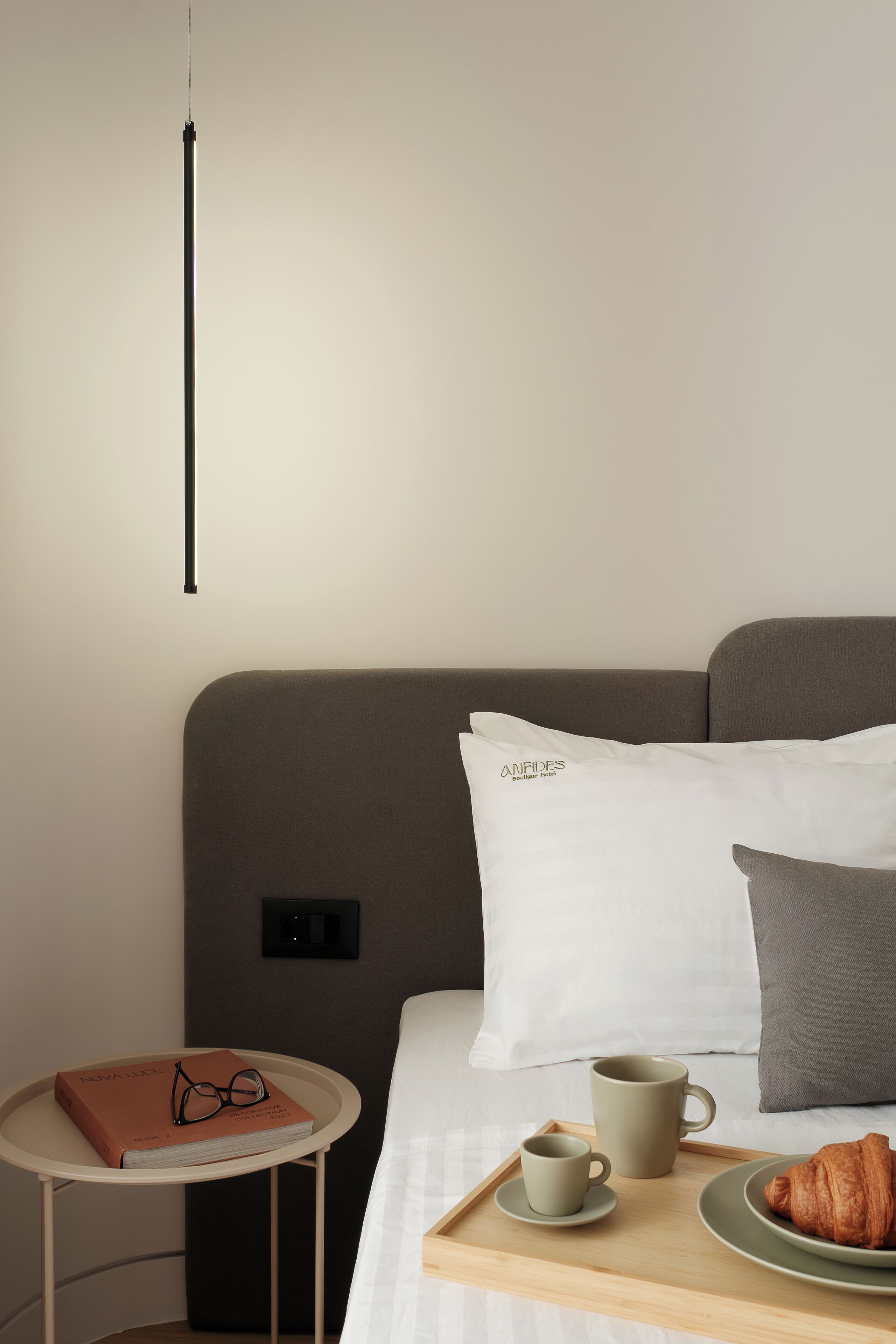A neatly arranged hotel room with a bed, a bedside table with a book and glasses, a tray with a croissant, a mug, and a plate.