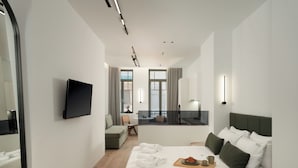 A modern hotel room with a bed, a TV, a sofa, a dining area, and a view of the city. - Anfides Boutique Hotel (Patras)