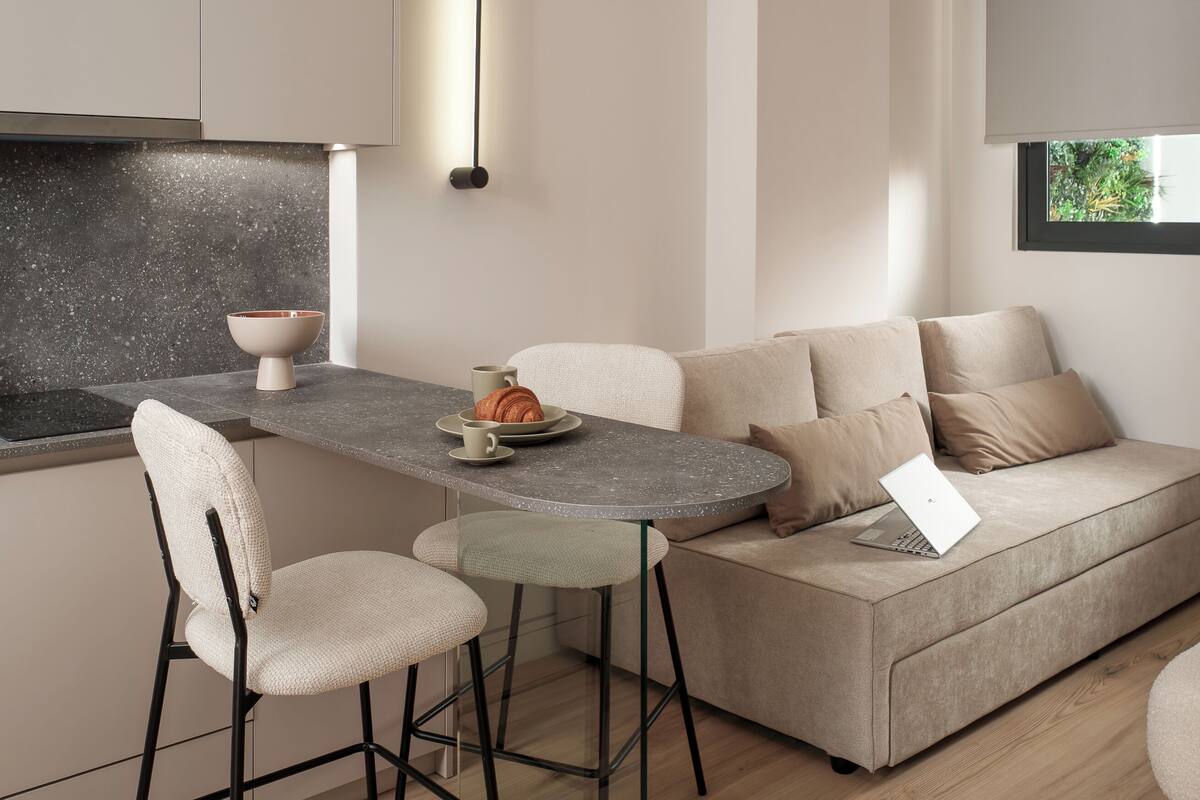 A modern kitchen with a dining area, featuring a round table, bar stools, and a sofa.