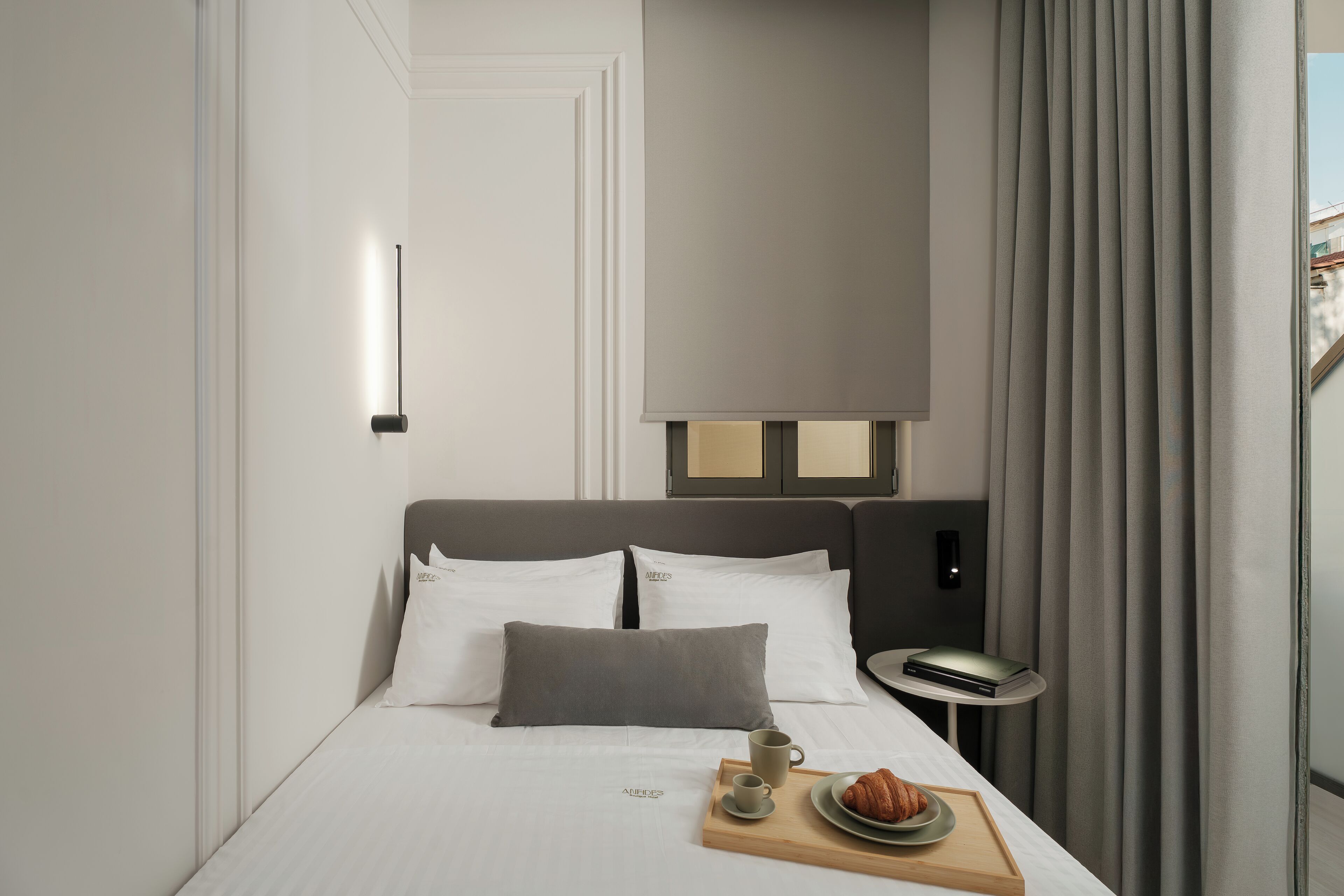 Habitación de hotel moderna con una cama bien hecha, una mesita de noche con lámpara y una bandeja de desayuno con croissants y café.