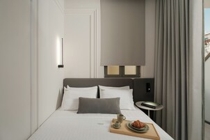 Habitación de hotel moderna con una cama bien hecha, una mesita de noche con lámpara y una bandeja de desayuno con croissants y café.