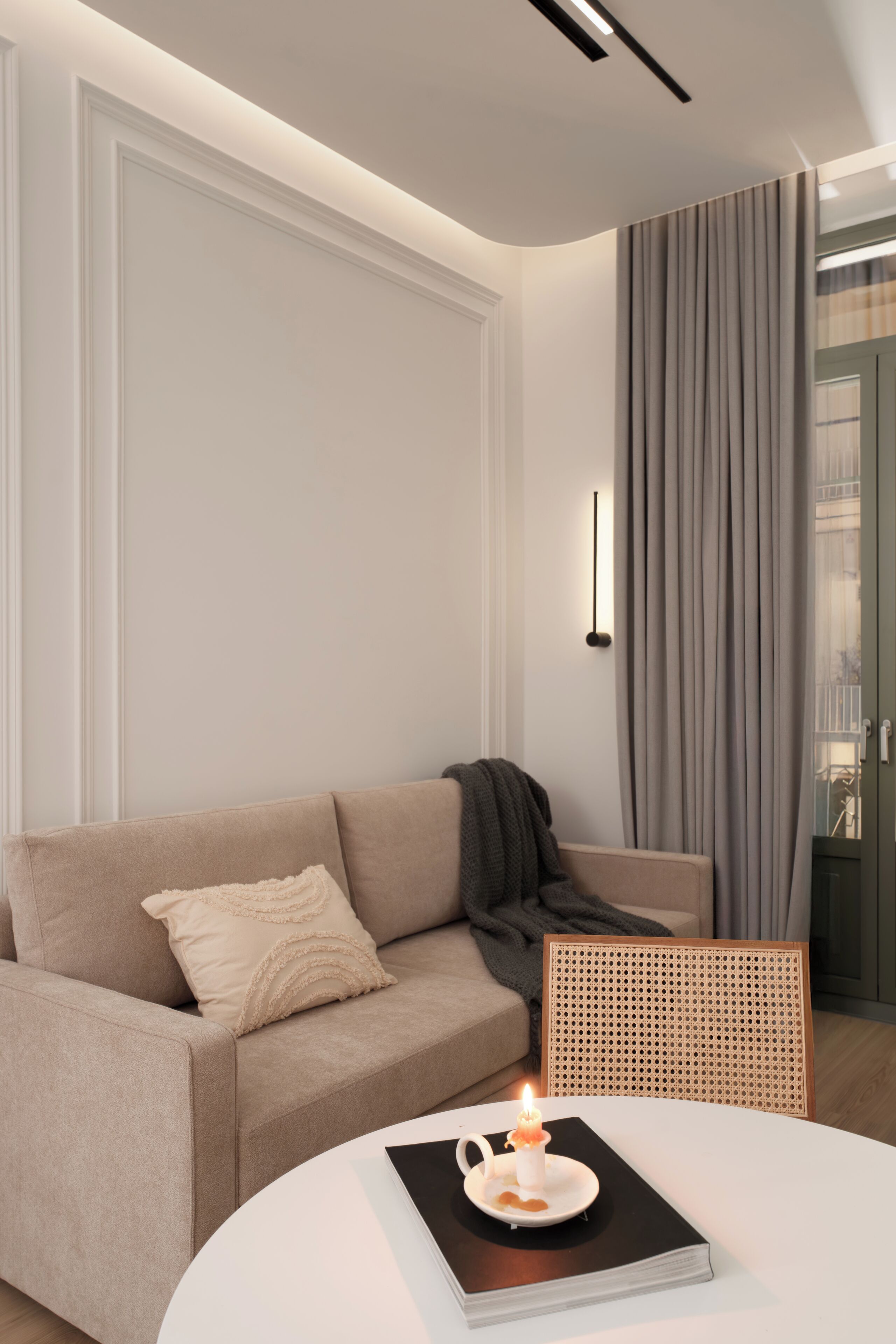 Ein modernes Wohnzimmer mit einem beigen Sofa, einem runden Couchtisch mit Buch und Kerze sowie einem Rattanstuhl.