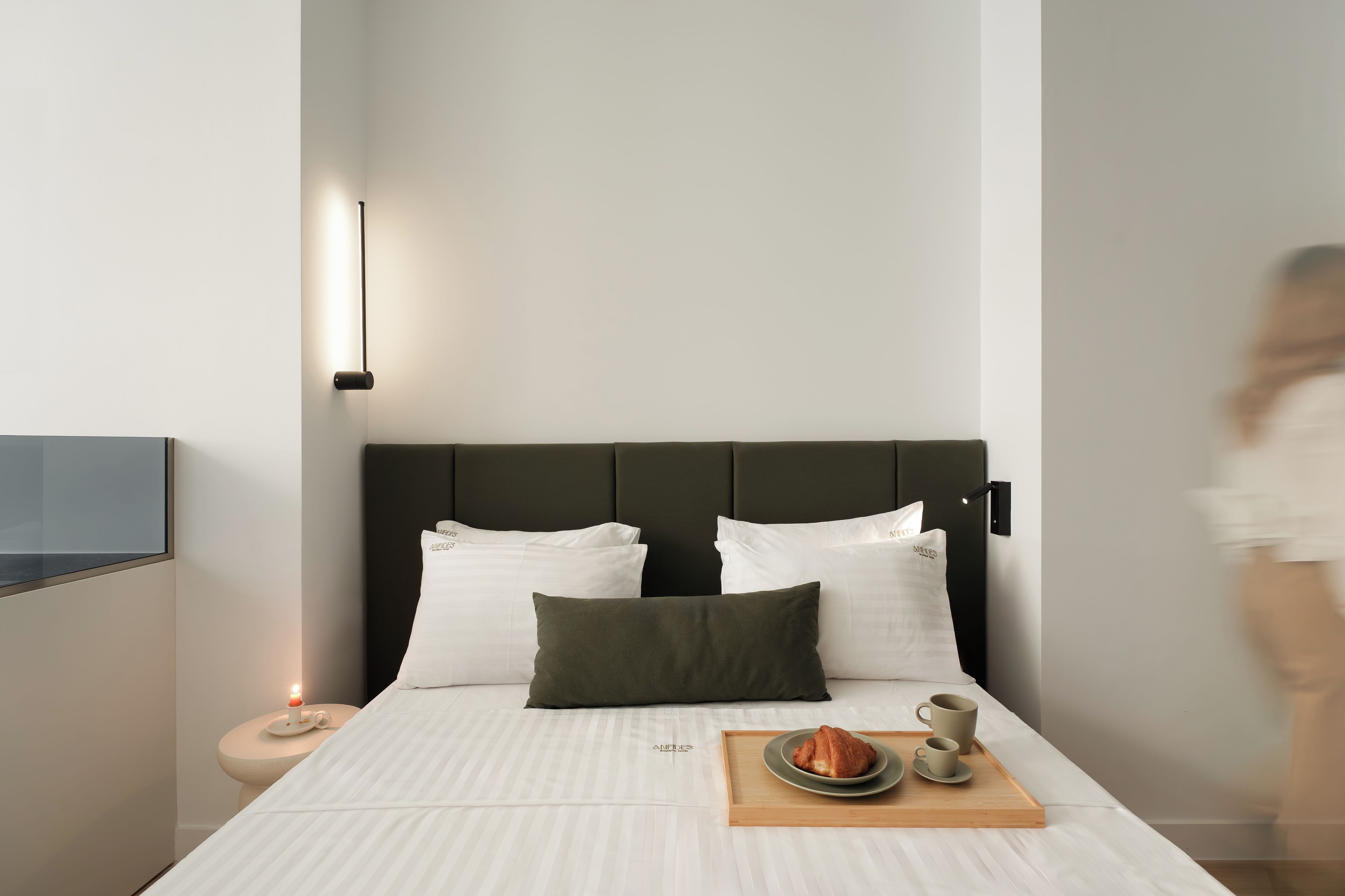 Ein modernes Hotelzimmer mit einem großen Bett, einem Nachttisch mit Lampe sowie einem Tablett mit Croissant und Kaffeetassen.