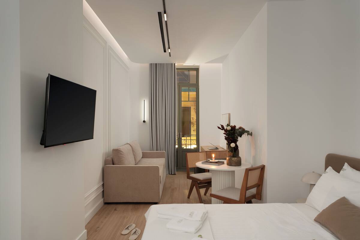 A modern hotel room with a flat-screen TV, a bed, a sofa, a desk with a chair, and a small table with a lamp.