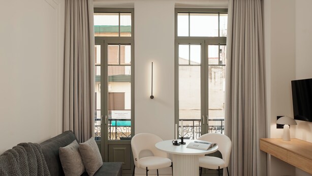 A modern living room with a grey sofa, a round table with two chairs, a TV on a wooden stand, and large windows with curtains. - Anfides Boutique Hotel (Patras)