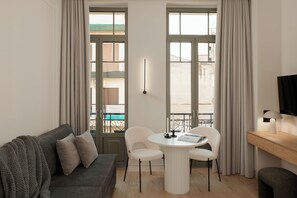 A modern living room with a grey sofa, a round table with two chairs, a TV on a wooden stand, and large windows with curtains. - Anfides Boutique Hotel (Patras)