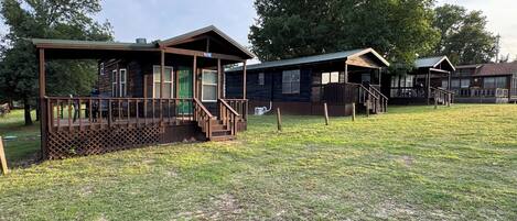A row of cabins with wooden decks and porches, surrounded by a grassy field and trees.