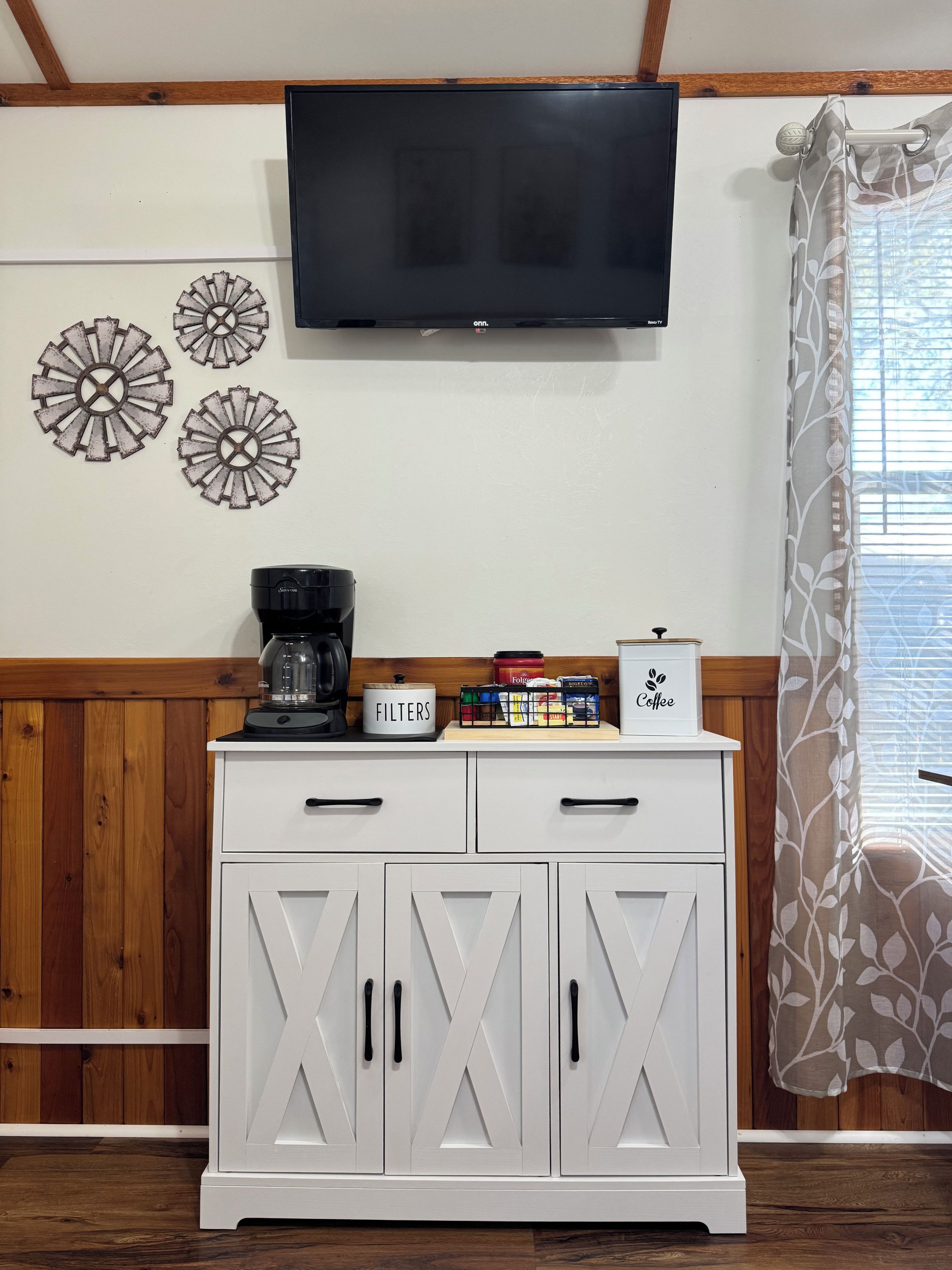 A room with a flat-screen TV mounted on the wall, a coffee maker on a cabinet, and decorative wall art.