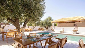 An outdoor dining area with wooden tables and chairs, a pool, and a pergola with hanging chairs.
