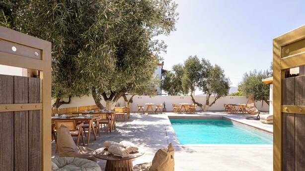 An outdoor pool area with wooden furniture, a fire pit, and olive trees.