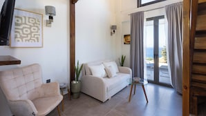 A living room with a sofa, a chair, a TV, and a balcony with curtains. - Eneos Residence - Koroni Poolside Jewel (QXW3+7GQ Koroni, Greece)