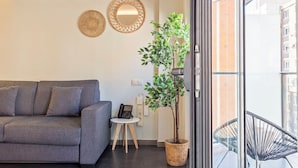 Una sala moderna con un sofá gris, una mesita, una planta en maceta y un balcón con una silla metálica.